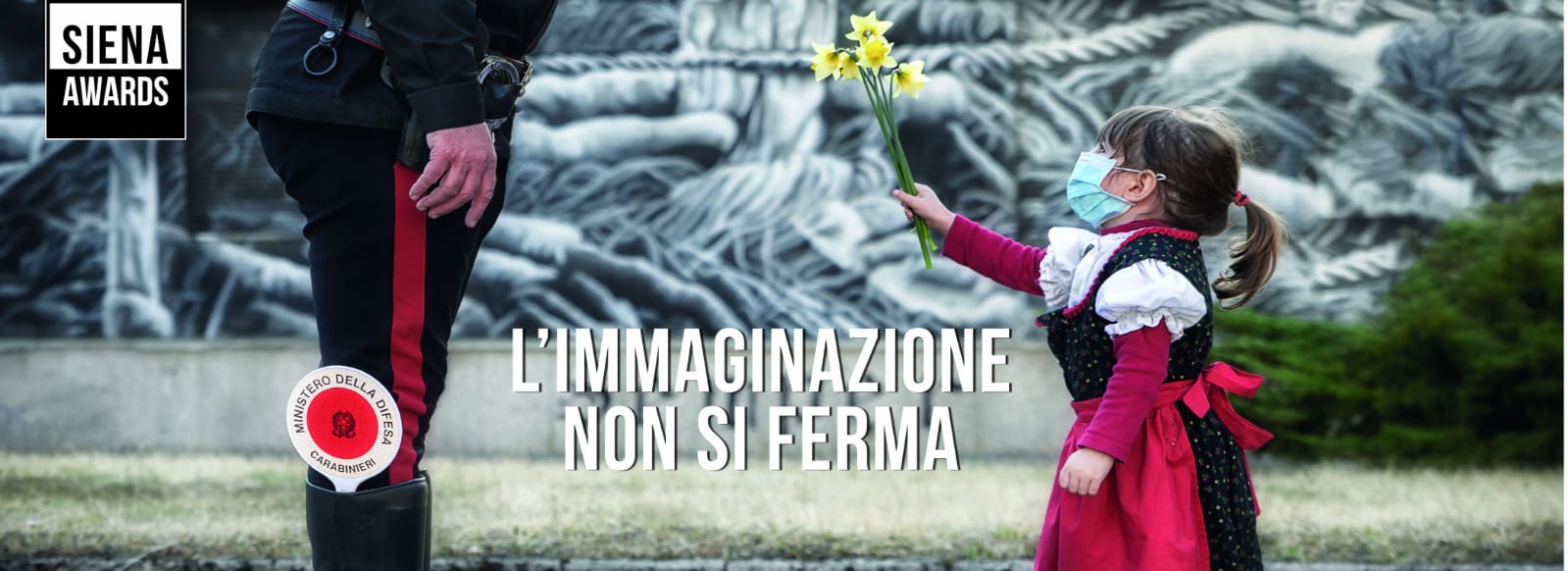L’immaginazione non si ferma. Foto di bimba con mascherina che dà fiore a Cc per Siena Award